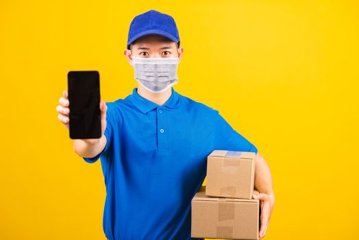 Asian young delivery worker man t-shirt and cap uniform wearing face mask protective show front black screen of smart mobile phone under coronavirus or COVID-19, studio shot isolated yellow background