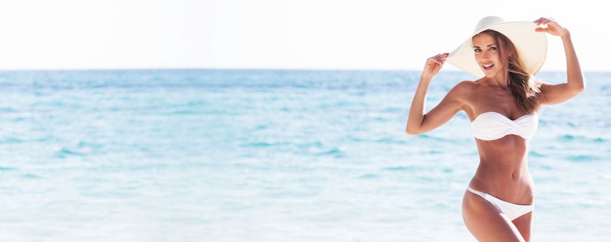 Smiling beautiful woman in white bikini and sun hat at sea beach