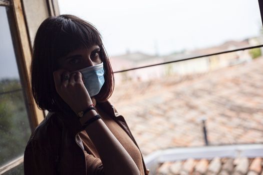 Girl with medical mask at window in her home during covid quarantine period