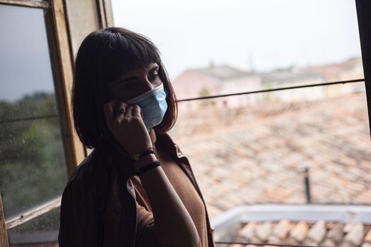 Girl with medical mask at window in her home during covid quarantine period