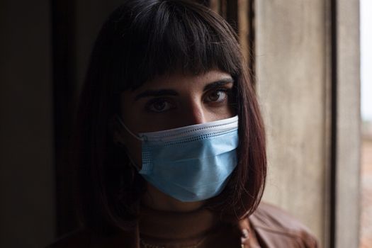 Girl with medical mask at window in her home during covid quarantine period