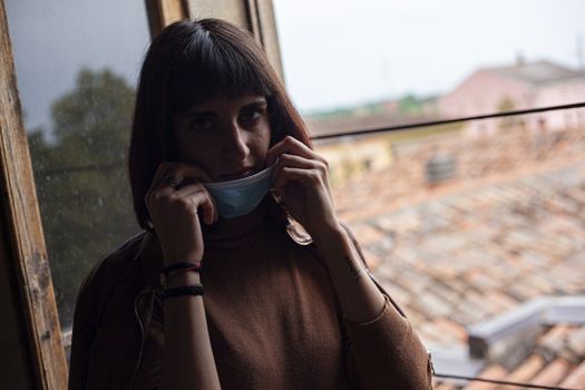 Girl with medical mask at window in her home during covid quarantine period