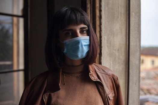 Girl with medical mask at window in her home during covid quarantine period