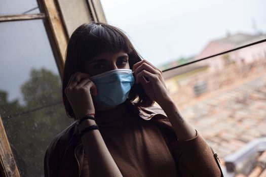 Girl with medical mask at window in her home during covid quarantine period