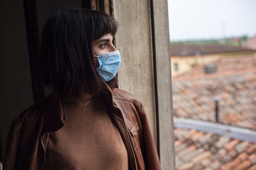 Girl with medical mask at window in her home during covid quarantine period