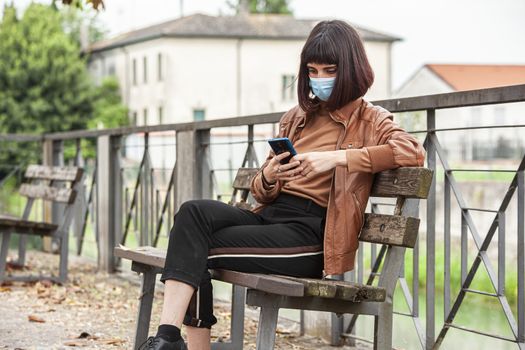 Portrait of a Girl with medical mask at park with smartphone during covid quarantine in Italy
