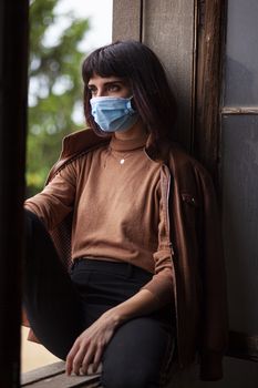 Girl with medical mask at window in her home during covid quarantine period