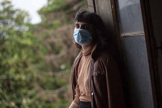 Girl with medical mask at window in her home during covid quarantine period