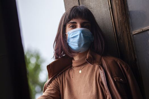 Girl with medical mask at window in her home during covid quarantine period