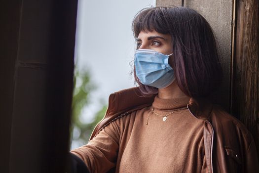 Girl with medical mask at window in her home during covid quarantine period