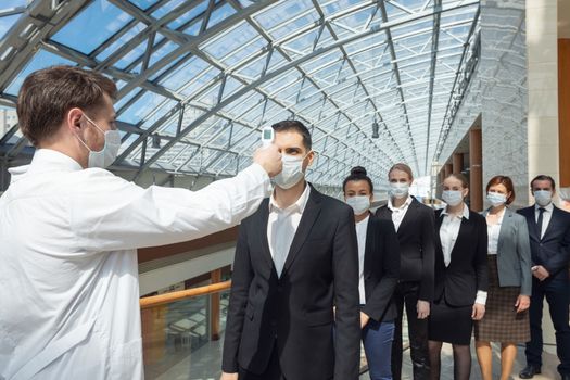 Doctor wearing protective surgical mask using infrared forehead thermometer (thermometer gun) to check body temperature for virus symptoms of business team waiting in a line