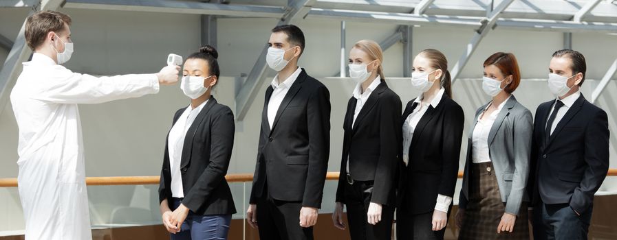 Doctor wearing protective surgical mask using infrared forehead thermometer (thermometer gun) to check body temperature for virus symptoms of business team waiting in a line