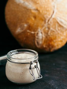 Sourdoug starter and wheat sourdough bread. Wheat sour dough starter in glass jar and delicious homemade round sourdough bread on black background. Home made sourdough bread making concept. Vertical