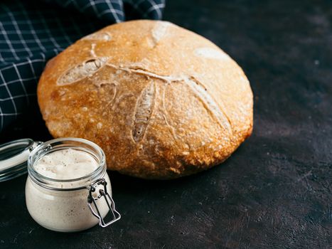 Sourdoug starter and wheat sourdough bread. Wheat sour dough starter in glass jar and delicious homemade round sourdough bread on black background, copy space. Home made sourdough bread making concept