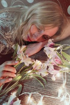 Portrait of a beautiful fashionable middle-aged woman with long blond hair, lying among white flowers and a garland of lights.