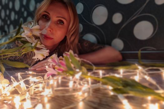 Portrait of a beautiful fashionable middle-aged woman with long blond hair, lying among white flowers and a garland of lights.