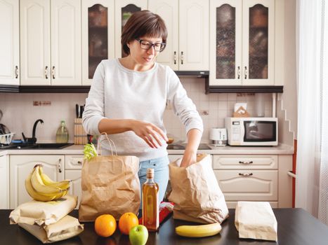 Woman sorts out purchases in the kitchen. Grocery delivery in paper bags. Subscription service from grocery store in conditions of quarantine because of coronavirus COVID19.