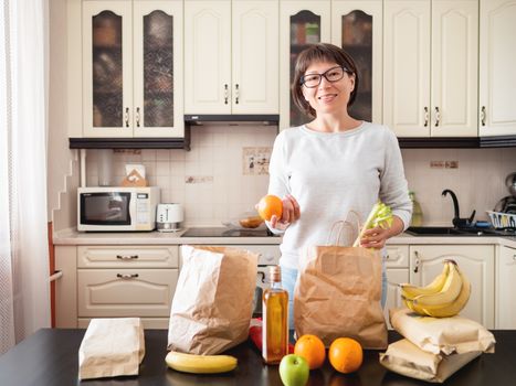 Woman sorts out purchases in the kitchen. Grocery delivery in paper bags. Subscription service from grocery store in conditions of quarantine because of coronavirus COVID19.