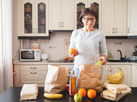 Woman sorts out purchases in the kitchen. Grocery delivery in paper bags. Subscription service from grocery store in conditions of quarantine because of coronavirus COVID19.