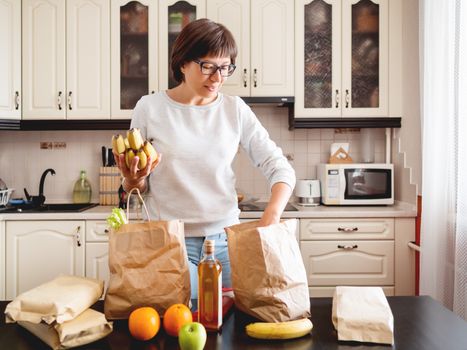 Woman sorts out purchases in the kitchen. Grocery delivery in paper bags. Subscription service from grocery store in conditions of quarantine because of coronavirus COVID19.