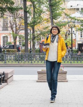 Take away coffee. Happy wide smiling women in bright yellow jacket holds paper cup with freshly brewed cappuccino. Hot beverage on cool autumn day.