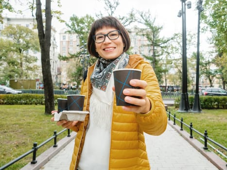 Take away coffee. Happy wide smiling women in bright yellow jacket holds paper cup with freshly brewed cappuccino. Hot beverage on cool autumn day.