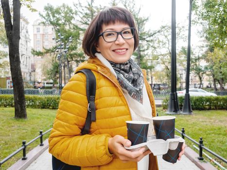 Take away coffee. Happy wide smiling women in bright yellow jacket holds paper cup with freshly brewed cappuccino. Hot beverage on cool autumn day.