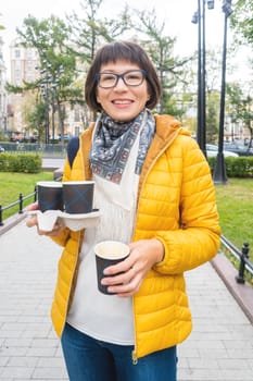 Take away coffee. Happy wide smiling women in bright yellow jacket holds paper cup with freshly brewed cappuccino. Hot beverage on cool autumn day.