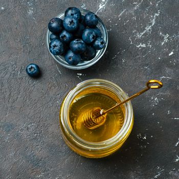 Healthy food ingredients - blueberries and honey. Glass jar with honey and dipper, fresh blueberries in small bowl over dark background. Top view or flat lay. Square crop. Copy space