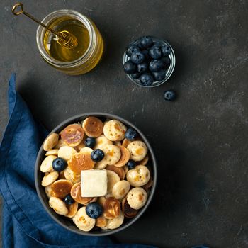 Trendy food - pancakes cereal served butter and blueberries. Heap of mini cereal pancakes in boul on dark background. Copy space right for text. Top view or flat lay. Square crop