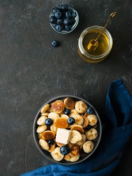 Trendy food - pancakes cereal served butter and blueberries. Heap of mini cereal pancakes in boul on dark background. Copy space right for text. Top view or flat lay. Vertical.