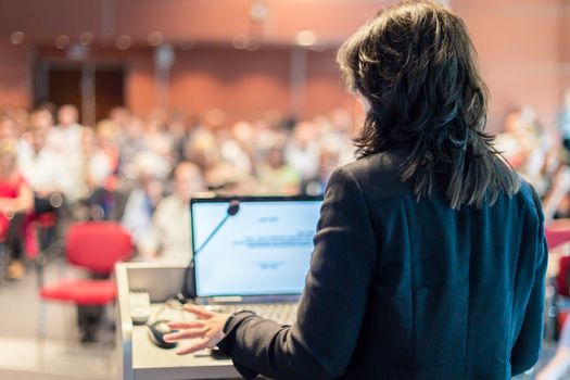 Female speaker giving a talk on corporate business conference. Unrecognizable people in audience at conference hall. Business and Entrepreneurship event.