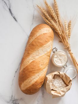 British White Bloomer or European sourdough Baton loaf bread on white marble background. Fresh loaf bread and glass jar with sourdough starter, flour in paper bag, ears. Top view. Copy space. Vertical