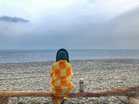 A lonely woman sits by the sea on a log, wrapped in a plaid, next to stands a thermos with tea.