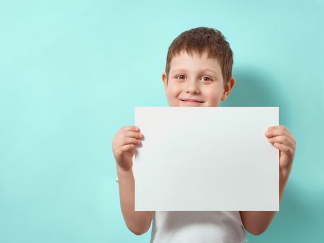 Four-year-old boy smiles and holds blank white paper sheet. Happy child on blue background with copy space for message, mock up