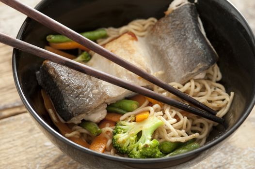 Asian noodles with fish fillet and fresh vegetables including broccoli and carrots served in a black bowl