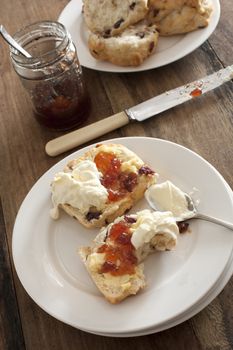 Tasty snack of a raisin scone or rock cake, fruit jam and whipped cream on a white plate with a bite missing in a high angle view