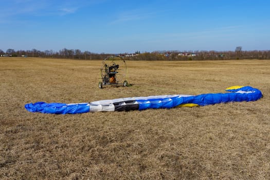 the parachute is spread on the dry grass on the field on a sunny day, in the background a paraglider