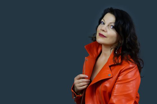 Portrait of a beautiful fashionable middle-aged woman in a bright orange leather jacket, posing on a dark gray background