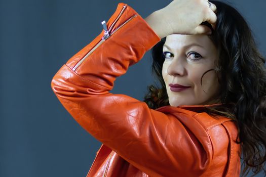 Portrait of a beautiful fashionable middle-aged woman in a bright orange leather jacket, posing on a dark gray background