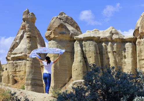 A young woman is holding a motley blue veil over her head, standing in the wind between huge and old rocks in the Cappadocia honey valley.