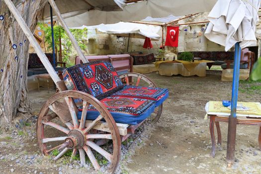 An old wooden cart on large wheels is fitted as a soft chair at a table under the tent in the old courtyard of a Turkish house.