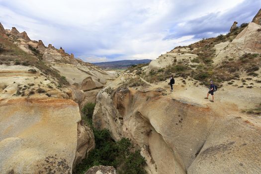 A young couple of tourists inspects a deep gorge standing on the edge of a canyon in Cappadocia against the background of a cloudy sky and mountain landscape.