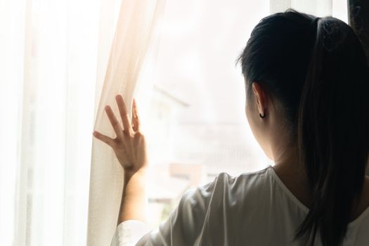 sad woman holding the curtains open to look out of a large light window at home, interior