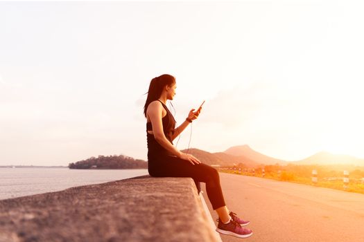 Side view of sport woman with earphones sitting on the wall after afternoon run. Female athlete taking a rest after outdoor workout