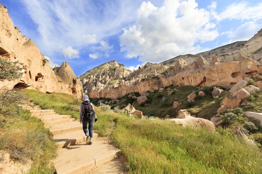 A young woman tourist walks along the road between huge, old, conical and weathered rocks in the Cappadocia honey valley under the bright day sun.