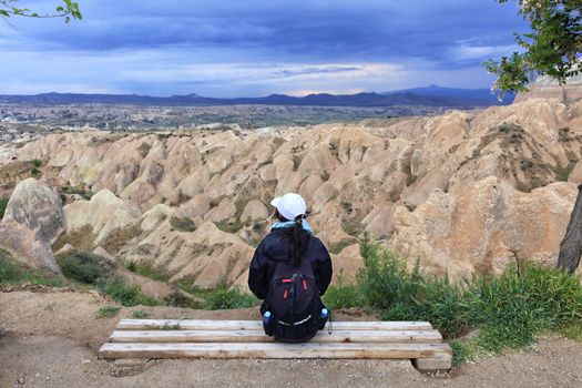 A young girl with a tourist backpack sits on a wooden bench and looks at the mountains, ravines and a stormy sky.