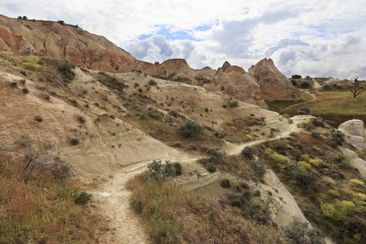 A mountain trail meanders along a road among a shrub between huge, old, conical and weathered rocks in the red valley of Cappadocia under a cloudy spring sky.