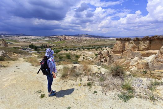 A young girl tourist with a backpack on her back is standing on the edge of a cliff in Cappadocia and admires the surrounding space against the background of a blue stormy sky and mountain scenery.