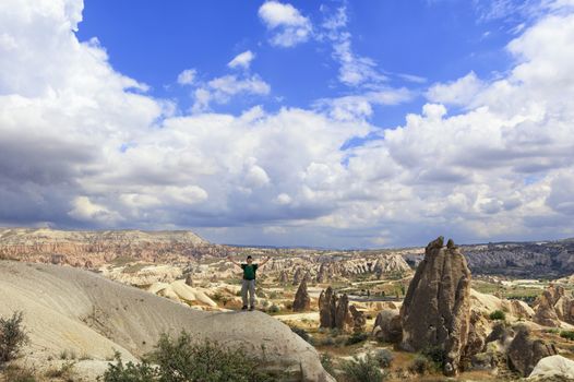 A young man in a green T-shirt and backpack on his back is standing on top of a hill in Cappadocia and looking up at the blue cloudy sky with his arms outstretched.